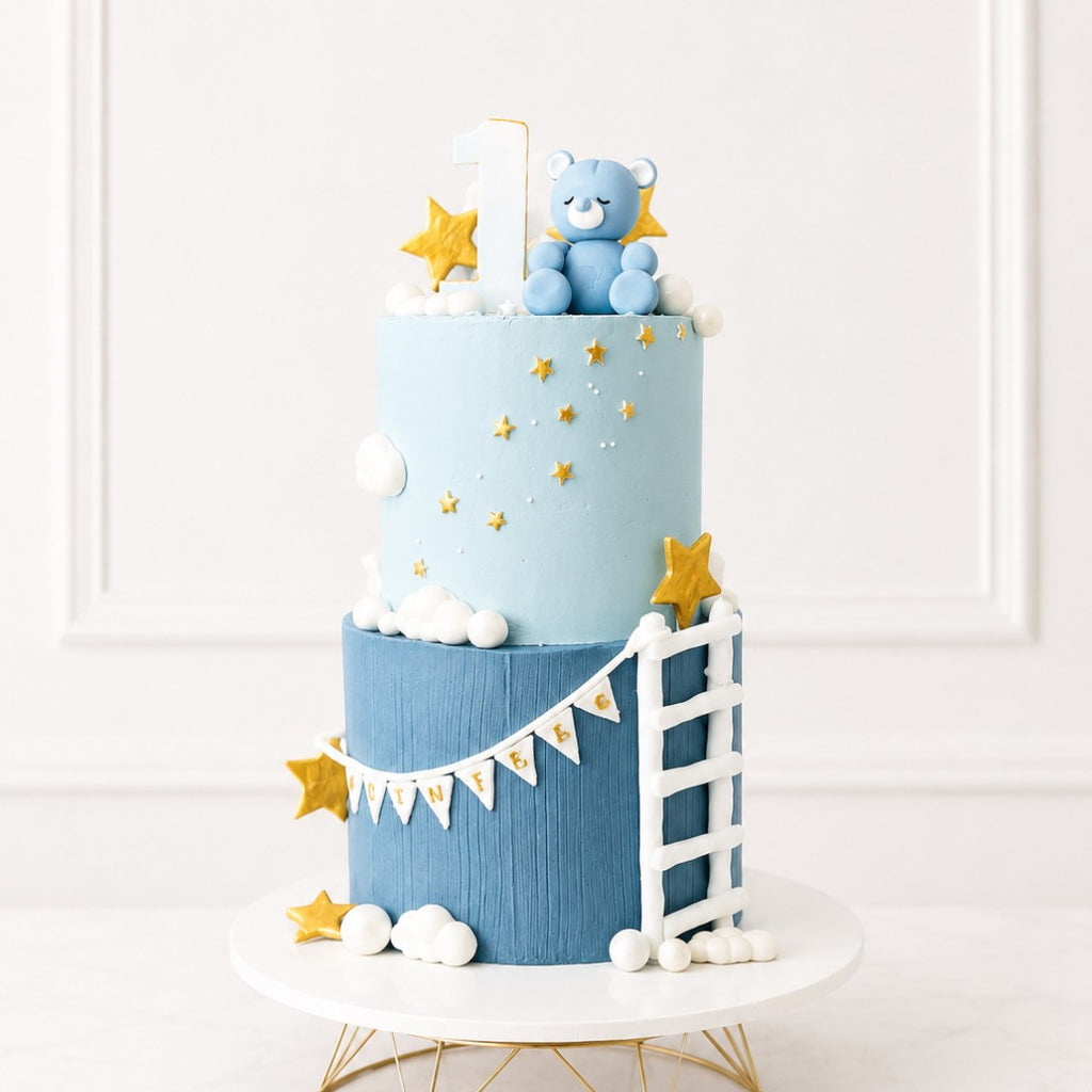 Two-tiered cake with blue and white color scheme, featuring a teddy bear and star decorations on a white background.