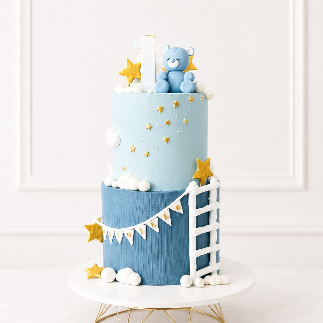 Two-tiered cake with blue and white color scheme, featuring a teddy bear and star decorations on a white background.
